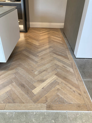 Wooden floor with herringbone pattern in a room with white walls and gray tiles.